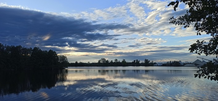 Baggersee im Abendlicht, eine dicke Wolke im Westen verdeckt die Sonne und wirft einen Schatten in unterschiedlichen Helligkeitsnuancen auf den See. Rechts oben von der Sonne angestrahlt weiße Wolken, die sich im See spiegeln.