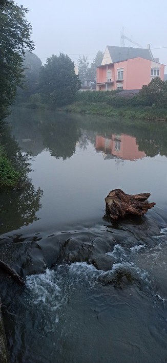 Rotes Haus spugelz sich im Fluss, der Morgennebel ist noch nicht ganz verschwunden. Vorne im Fluss fällt das Wasser über eine Kante