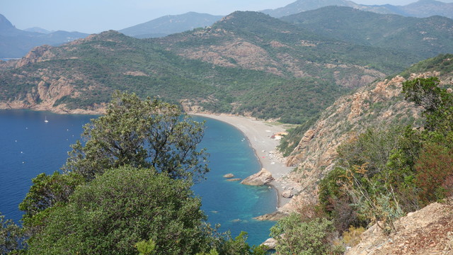 Blick von einer Straße oben auf einem Berg herunter zum Meer, das türkis schimmert. Rechts fallen die Berge steil zu einem kleinen Sandstrand ab, der sich ca. 200 lang ist. Wellengang keiner, ein paar Boote for dem Ufer. Dahinter Berge Korsicas begrünt und mit vielen Bäumen bis in den Horizont.