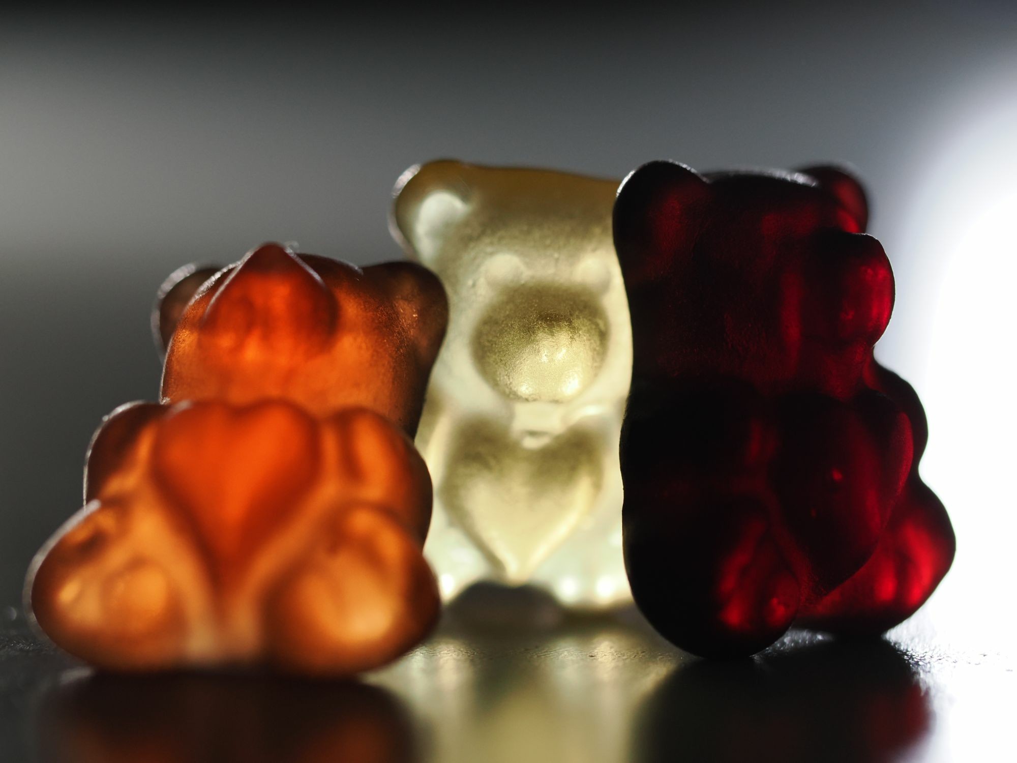 Drei Gummibärchen mit einem Herz auf dem Bauch sitzen aneinander gelehnt auf einer glatten Fläche. Sie sind von hinten beleuchtet, das linke ist hellrot, das mittlere weiß, das rechte ein sehr dunkles rot.