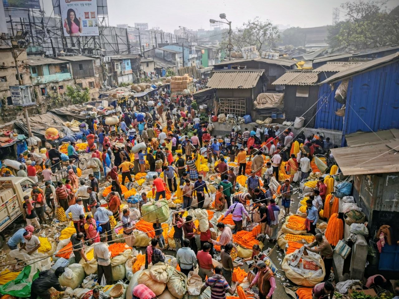 Foto eines Marktes, von oben fotografiert. Zwischen schäbigen Hütten und Häusern ein Gedränge aus Menschen, Säcken und Blüten und Girlanden in Rot und Orange. Im Hintergrund sind Hochhäuser zu sehen.