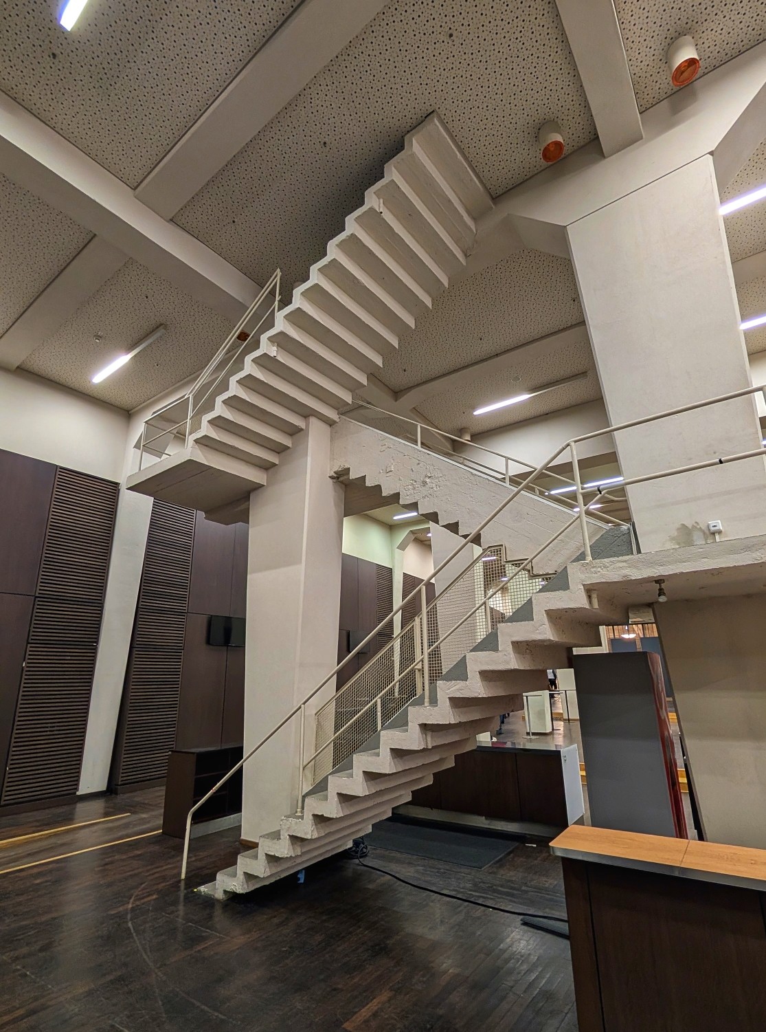 Das Bild zeigt einen Innenraum mit einer Wendeltreppe als zentrales Element. Die Treppe ist weiß und hat Metallgeländer. Sie führt von unten nach oben und biegt sich um einen vertikalen Schacht. Auf beiden Seiten der Treppe befinden sich Wände, die auf der linken Seite dunkelbraun und auf der rechten Seite hellgrau sind. Im Hintergrund ist ein Teil des Gebäudedachs zu sehen. Auf der rechten Seite befindet sich ein niedriger Schalterkasten und ein kleines dunkles Schreibtischarrangement. Die Treppe führt direkt auf eine Decke zu und kann nicht bis oben begangen werden.