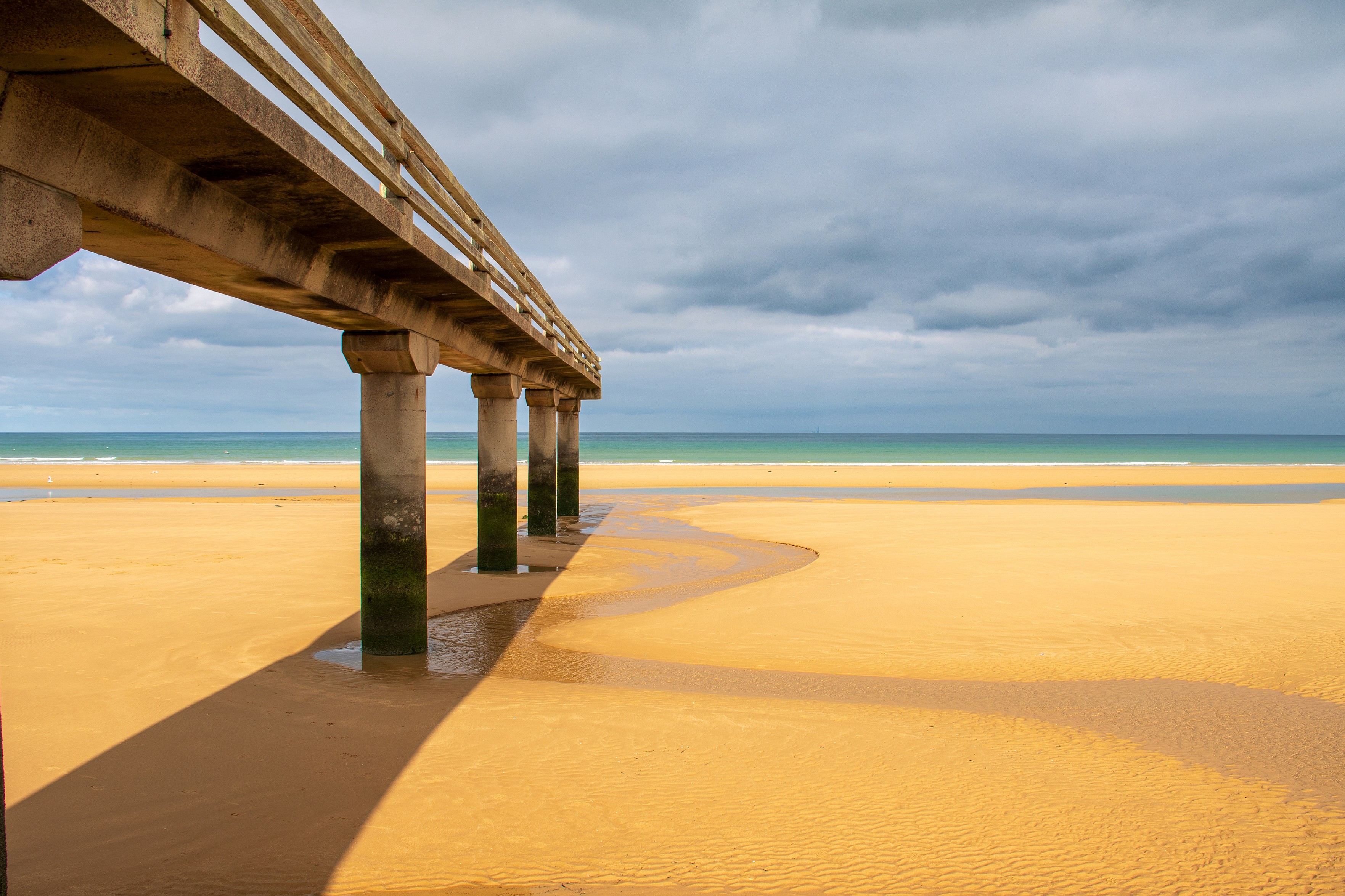 Das Bild zeigt einen Pier, der sich über einen gelben Sandstrand erstreckt. Der Pier hat Betonpfeiler, von denen einige mit grünem Bewuchs bedeckt sind. Die Pfeiler werfen lange, dunkle Schatten auf den Sand. Im Hintergrund sieht man den Ozean unter einem bewölkten Himmel. Die Farben im Bild sind gedämpft, mit einem Kontrast zwischen dem warmen Gelb des Sandes und dem kühlen Blau und Grau des Himmels und des Ozeans.