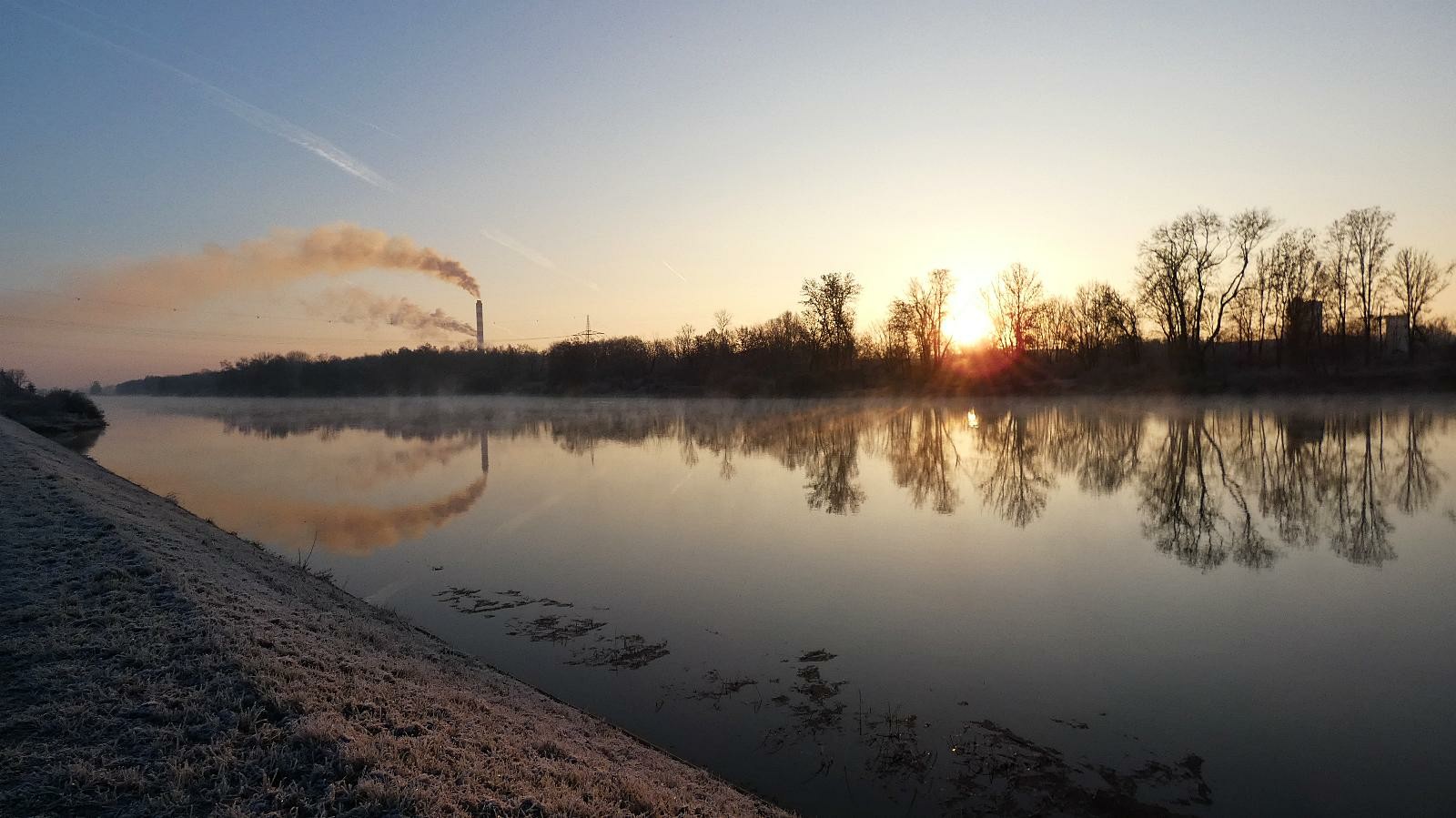 Morgentlicher Blick auf die Donau,in ihr spiegeln sich das andere Ufer mit Bäumen,die Sonne,und ein paar Schornsteine mit Rauch.