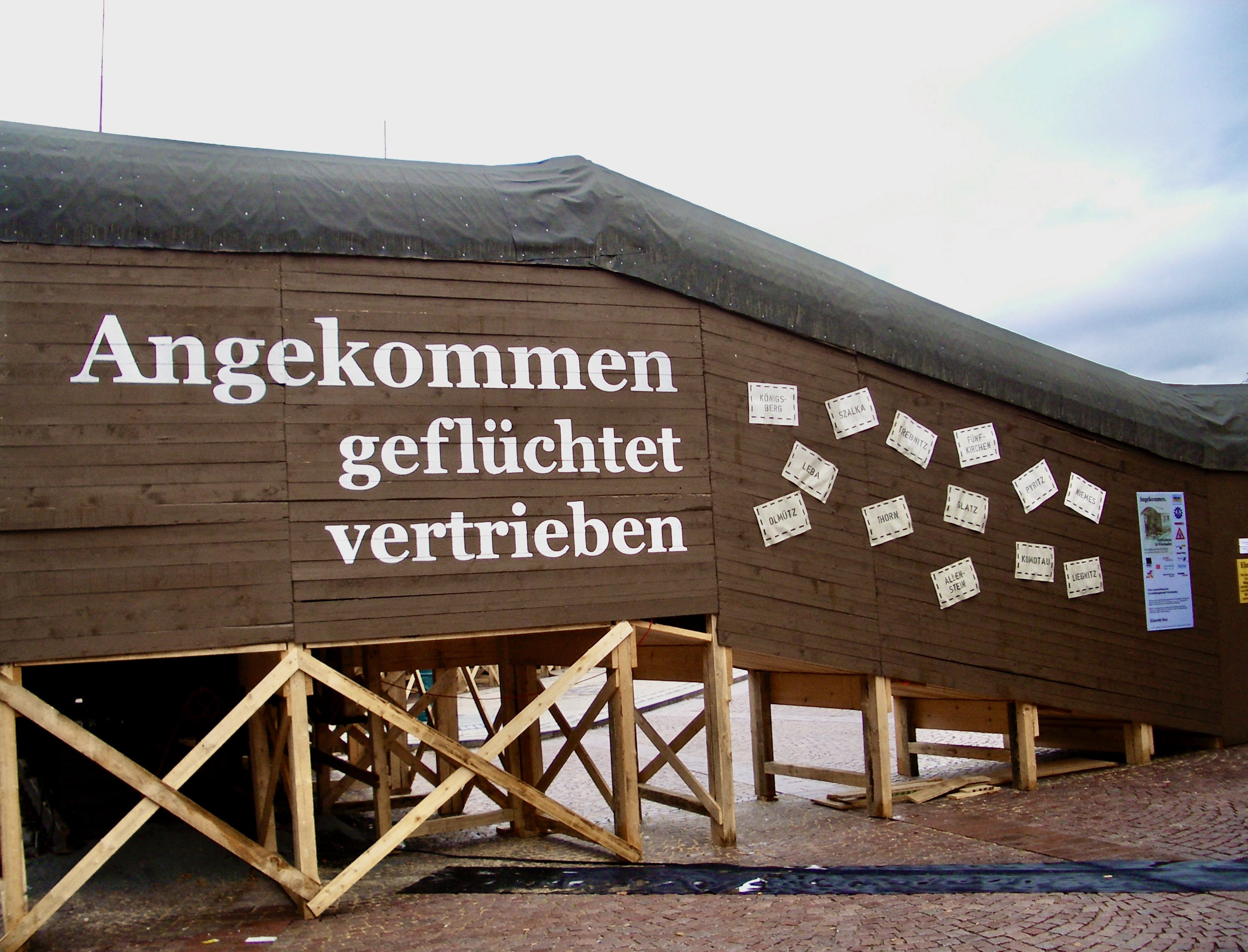 Das Bild zeigt die hölzerne Außenseite eines Gebäudes oder einer Struktur mit einem schrägen Dach. An der Holzfassade befinden sich große weiße Buchstaben, die den Text "Angekommen geflüchtet vertrieben" bilden. Neben dem Text sind zahlreiche kleine, weiße, rechteckige Zettel mit Ortsnamen aus den ehemaligen deutschen Ostgebieten an der Wand befestigt. Das Gebäude wird von einer Konstruktion aus Holzbalken gestützt. Im Vordergrund ist ein roter Untergrund zu sehen, möglicherweise eine Art Bodenbelag oder Pflaster