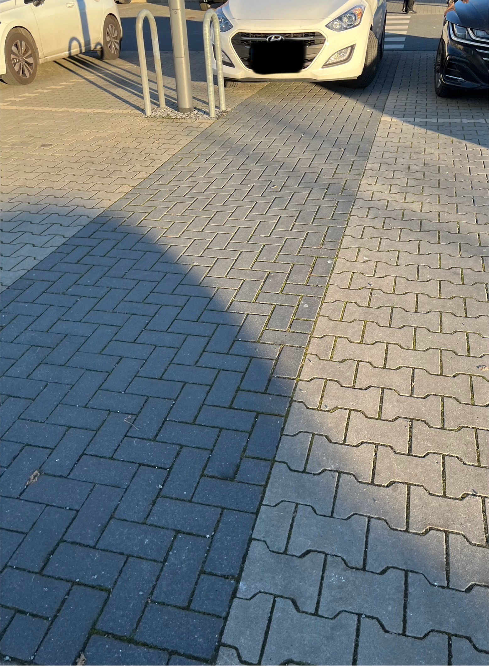 Ein Parkplatz. Ein Auto ( weißer Hyundai) parkt auf einem dunkel abgesetzten Streifen- dieser geht in einen Zebrastreifen über. Stellt also relativ offensichtlich einen Fußweg dar. Neben dem Auto ist auch direkt ein Parkplatz auf einer helleren Fläche frei. Das Auto steht da zu auch drauf.