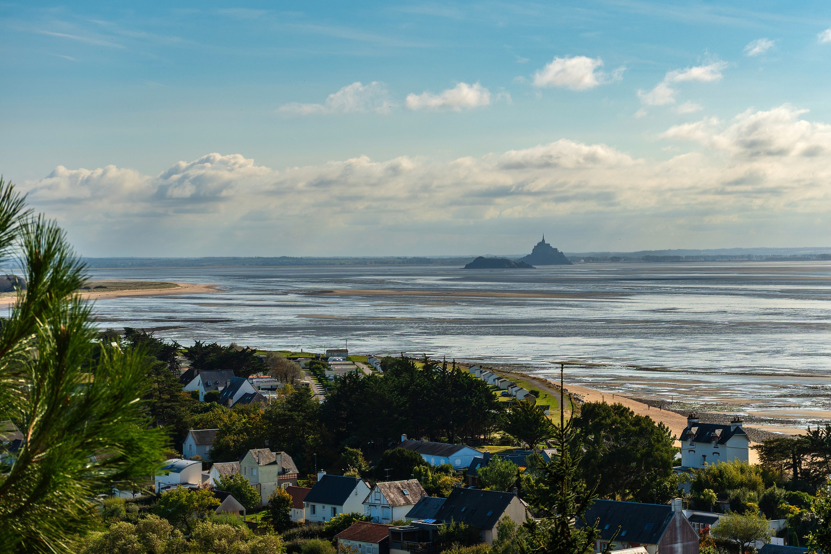 Das Bild zeigt eine Ansicht von Mont Saint-Michel, einer Gezeiteninsel in der Normandie, Frankreich. Im Vordergrund ist eine Ansicht einer Kรผstenstadt mit Hรคusern und Vegetation. Dazwischen befindet sich ein weites Wattenmeer, das bei Ebbe freiliegt. Im Hintergrund erhebt sich Mont Saint-Michel mit seiner charakteristischen Silhouette und der Spitze der Abtei. Der Himmel ist blau mit einigen Wolken, was auf einen klaren Tag hindeutet.