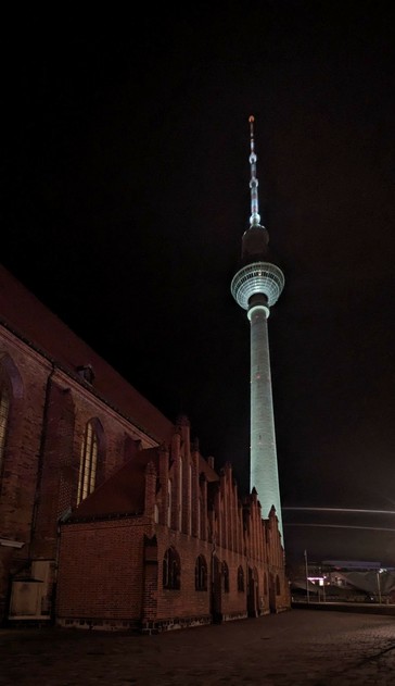 Dunkel, beleuchteter Alexander Funkturm ragt in den Himmel und Teile der Backsteinkirche St Marien links