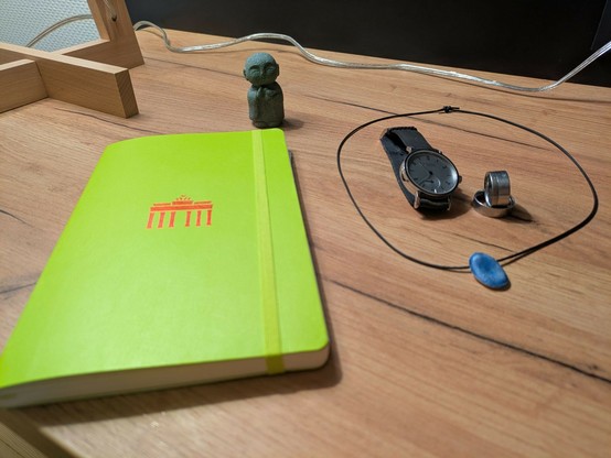 ein hellgrünes Notizbuch mit einem roten Symbol vom Brandenburger Tor drauf liegt auf einem Holztisch. ein sehr kleiner Jizo Buddha mit gefalteten Händen steht dahinter. Eine Uhr, 2 Edelstahlringe liegen innerhalb einer ledernen Halskette mit blauem Anhänger