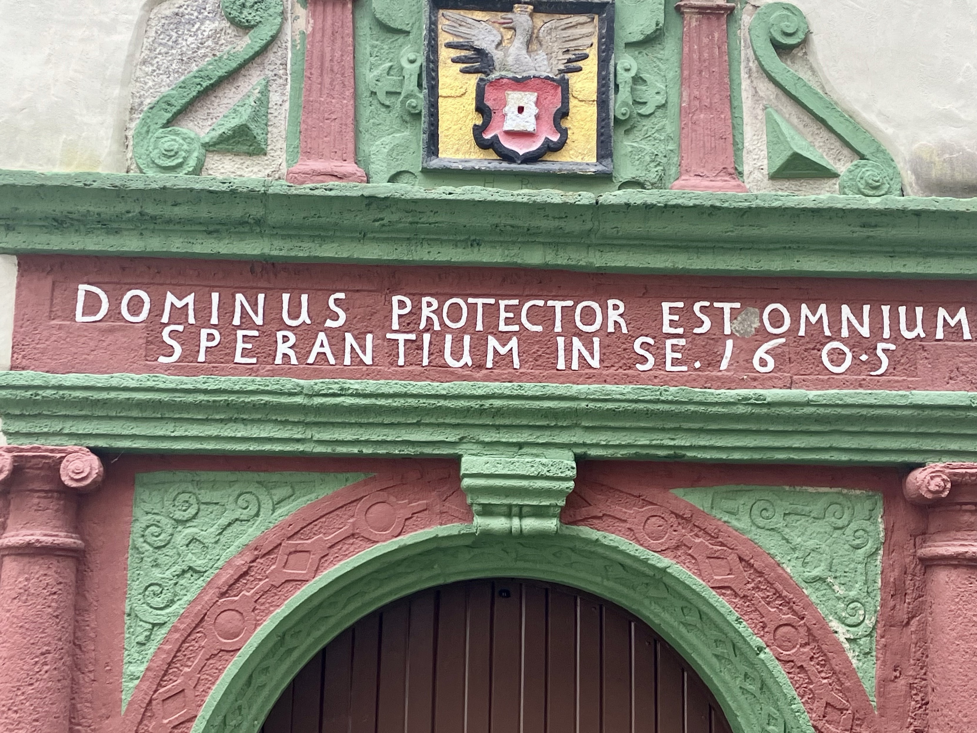 Ein dekorativer architektonischer Eingang mit einem rötlich-braunen Torbogen mit grünen Akzenten. Über dem Bogen befindet sich ein geschnitzter Schild, der mit einem Adler und einem Wappen geschmückt ist. Lateinischer Text mit der Lesung "DOMINUS PROTECTOR EST OMNIUM SPERANTIUM