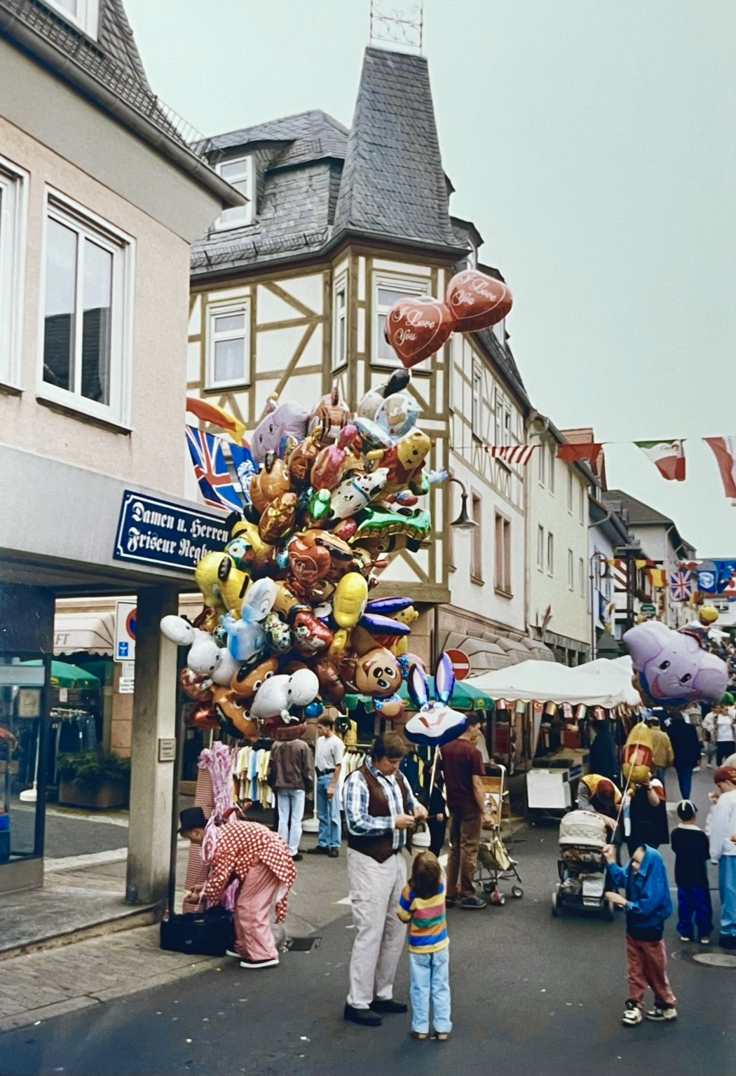 Ein Ballonverkäufer, der eine Menge bunter Luftballons in der Hand hält, verkauft einen der Ballons an ein Kind. Dies geschieht einer Fußgängerzone, umgeben von Häusern.