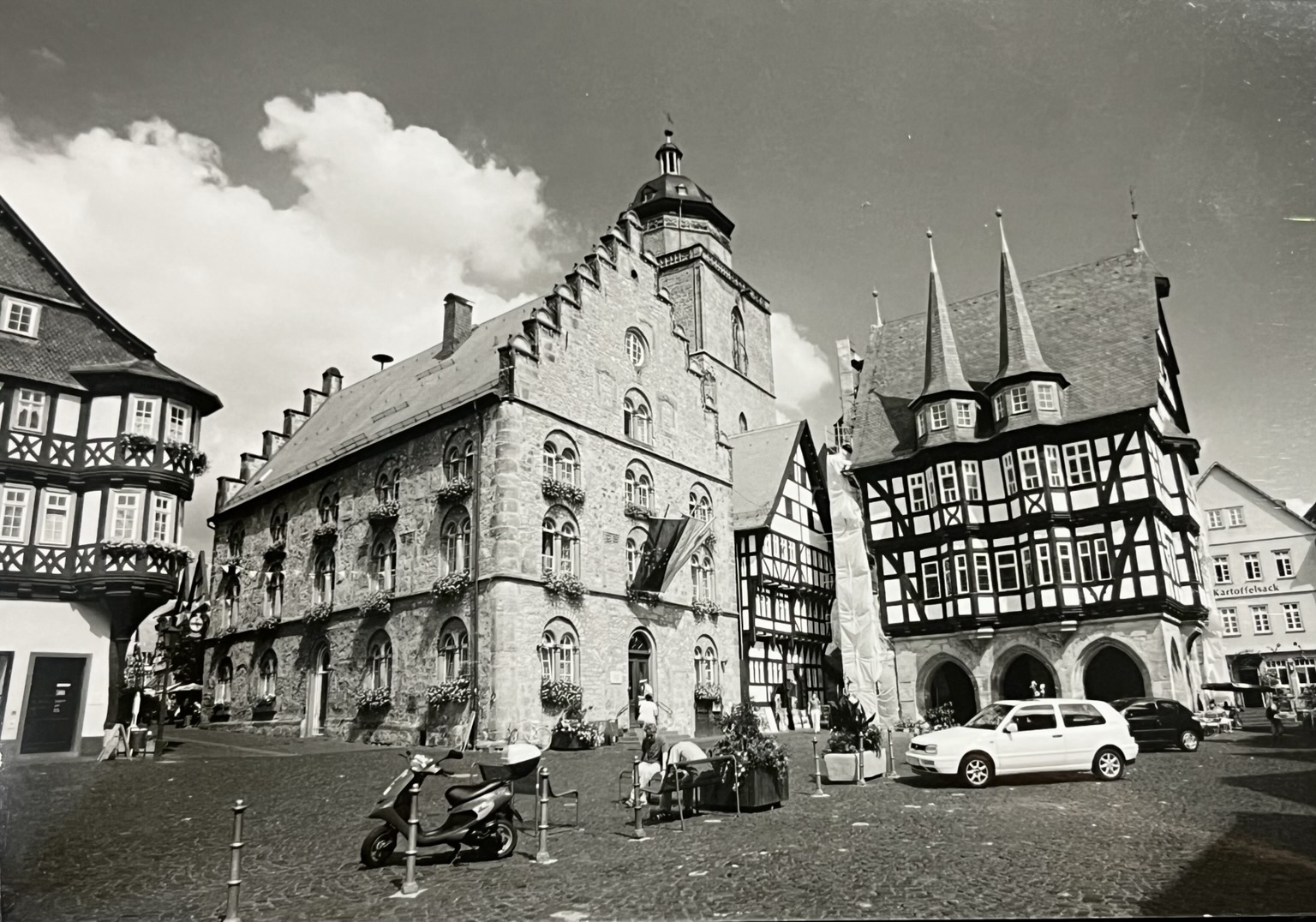 Mittelalterliche Häuser. Rechts ein Fachwerkhaus mit kleinen Türmchen, links ein aus Stein gemauertes Haus. Zwischen beiden sieht man einen Kirchturm. Vor dem Fachwerkhaus parkt ein weißes Auto. Schwarz-weiß Bild.