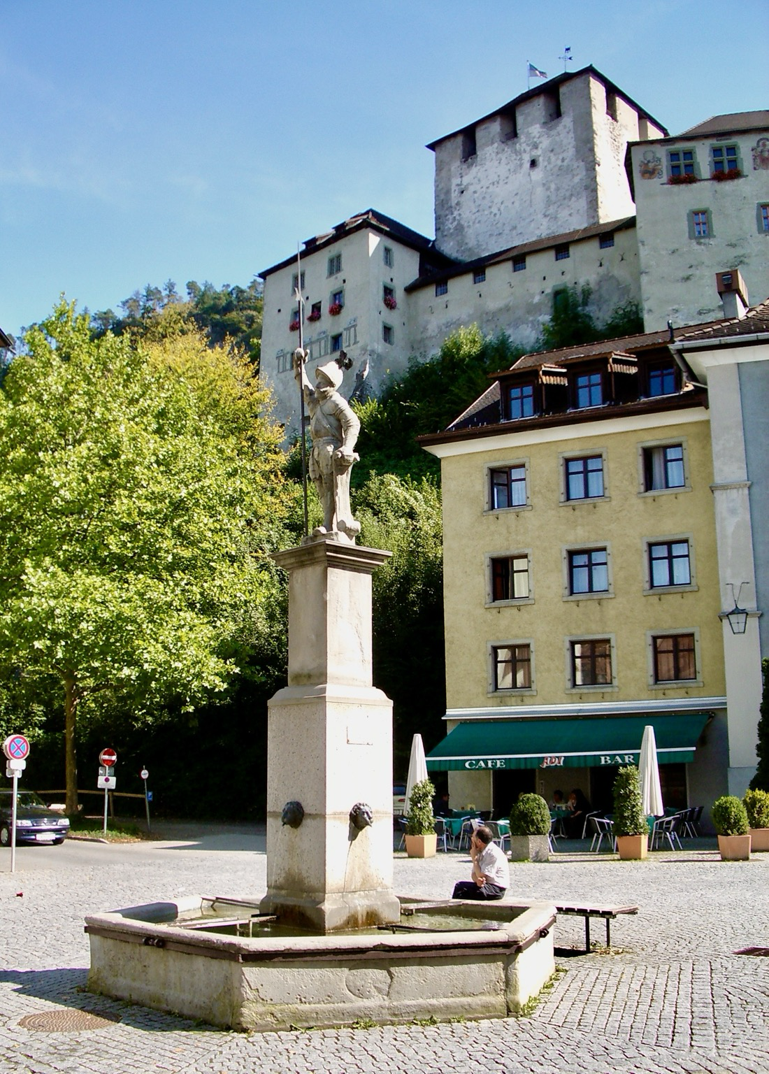 Ein Springbrunnen, in dem auf einer hohen Säule eine Statue steht. Im Hintergrund links ein gelbes Haus, und rechts ein großer grüner Baum, alles überragt von der Schleppenburg