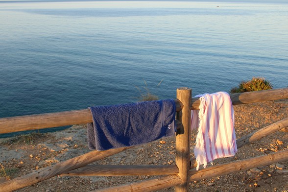 ein dunkelblaues und hellblaues Badehandtuch hängen über einen aus 10 cm dicken Rundhölzern bestehenden Zaun, der an der Klippenkante steht. Die Handtücher werden von der ersten Morgensonne leicht orange angeleuchtet dahinter das Meer, dunkelblau, ruhig und ohne Wellengang. 