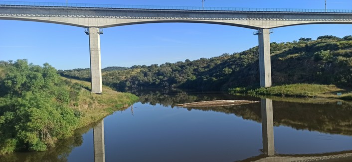einen neue Eisenbahnbrücke aus Beton und oben mit Geländer versehen steht mit zwei Pfeilern am Uferrand eines 100 Meter breiten Sees und überspannt diesen. Im Wasser ist das steinerne Geländer der alten Rundbogenbrücke zu sehen, die wegen dem hohen Wasserstand noch fast überflutet ist. Szene ist in das helle Morgenlicht der Sonne getaucht, Himmel strahlend blau, die Eisenbahnbrücke spiegelt sich mit den 25 Meter hohen Pfeilern im Wasser.