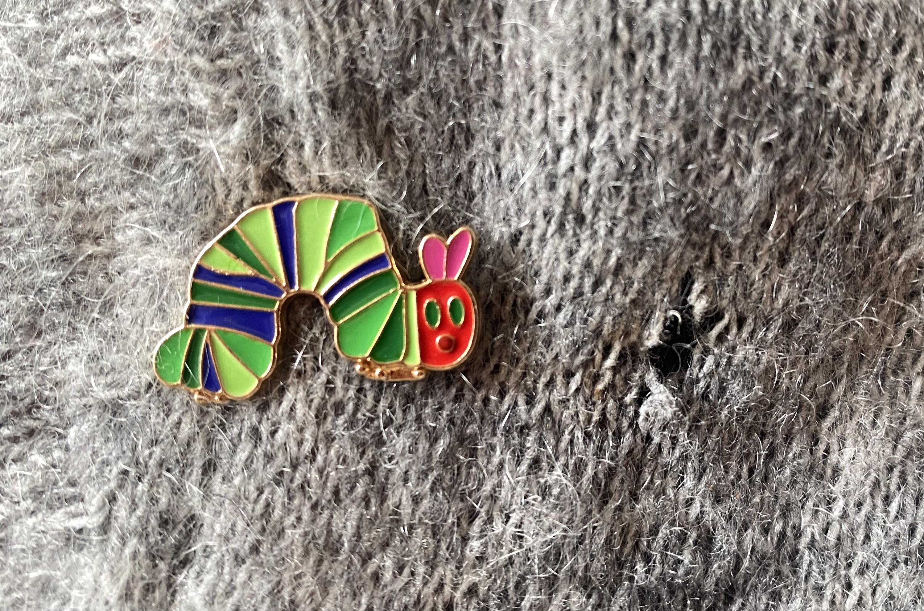 Nahaufnahme eines flauschigen Gestricks mit kleinem Loch. Daneben wurde das bunt emaillierte Pin der Raupe Nimmersatt platziert.