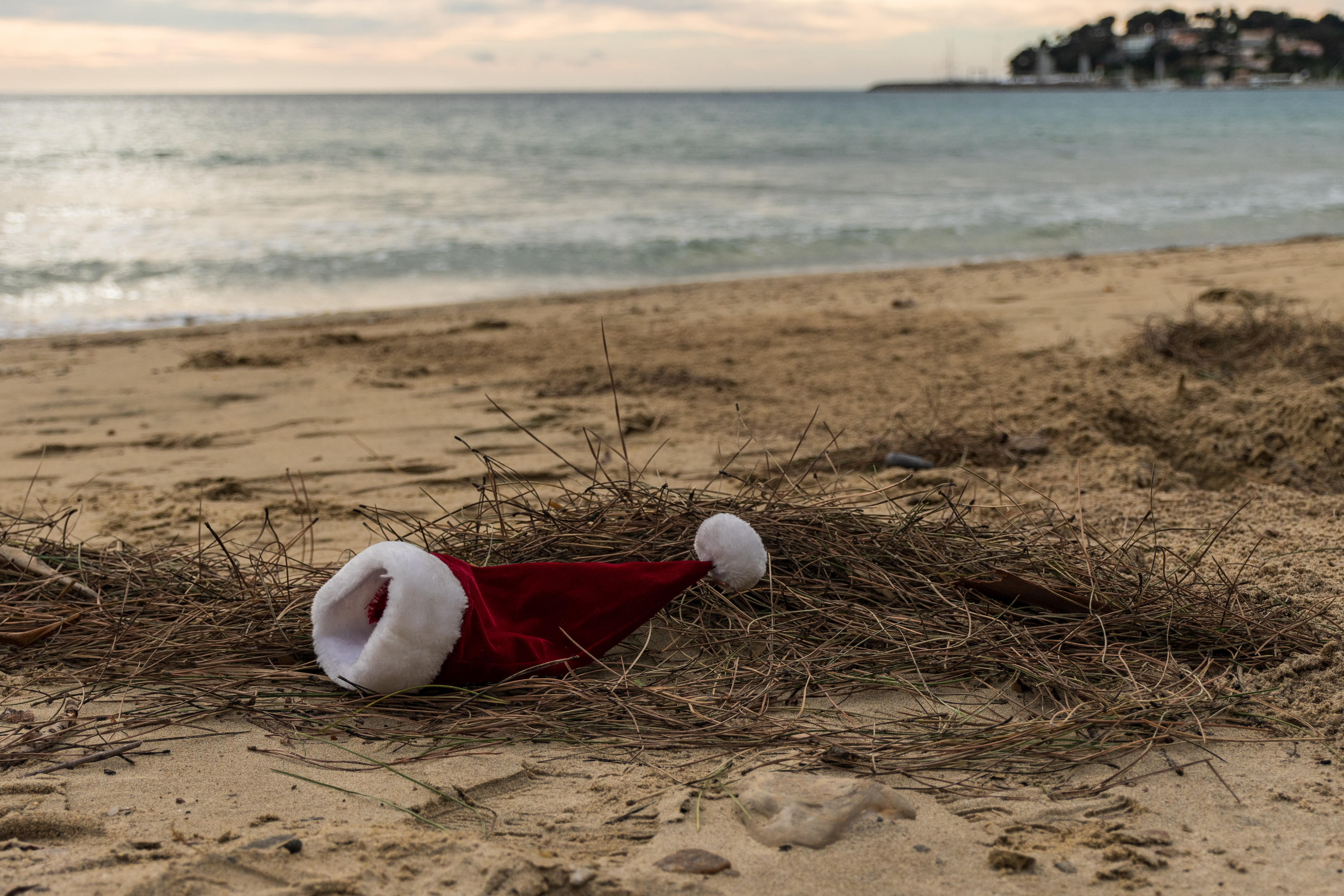 Das Bild zeigt eine rote Weihnachtsmütze mit weißem Rand und Bommel, die im Sand an einem Strand liegt. Im Hintergrund ist das Meer und ein bewölkter Himmel sichtbar.