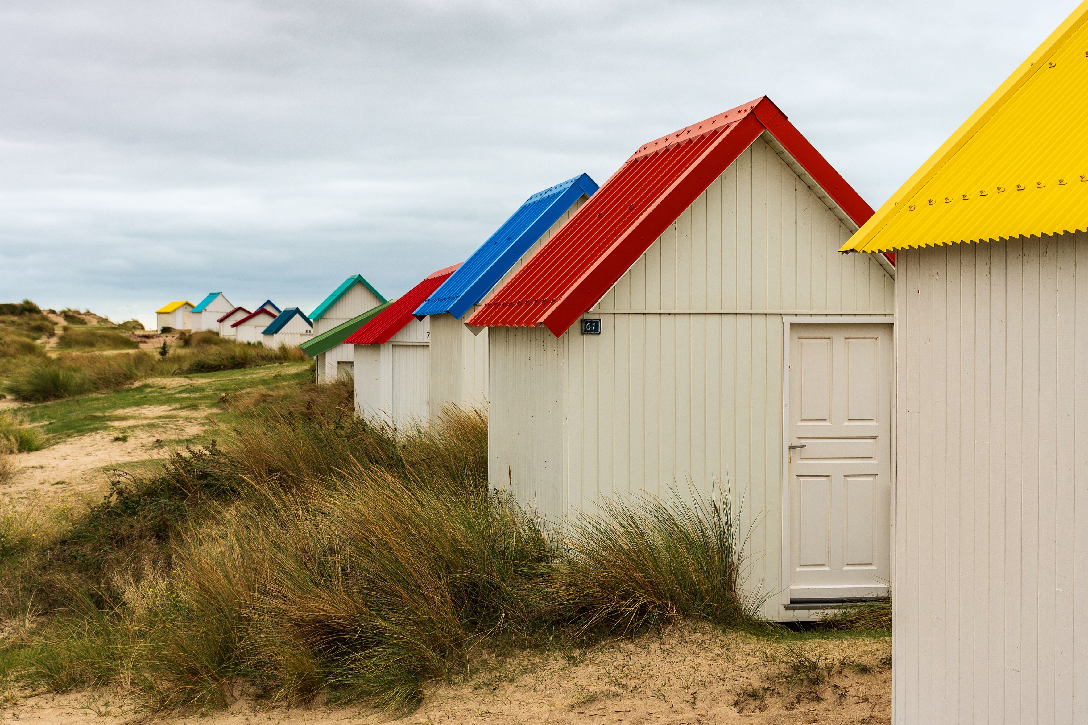 Das Bild zeigt eine Reihe von kleinen, weißen Strandhäusern mit bunten Dächern (gelb, rot, blau, grün). Die Häuschen stehen in einer Reihe auf Sanddünen, die mit Gras bewachsen sind. Der Himmel ist bewölkt.