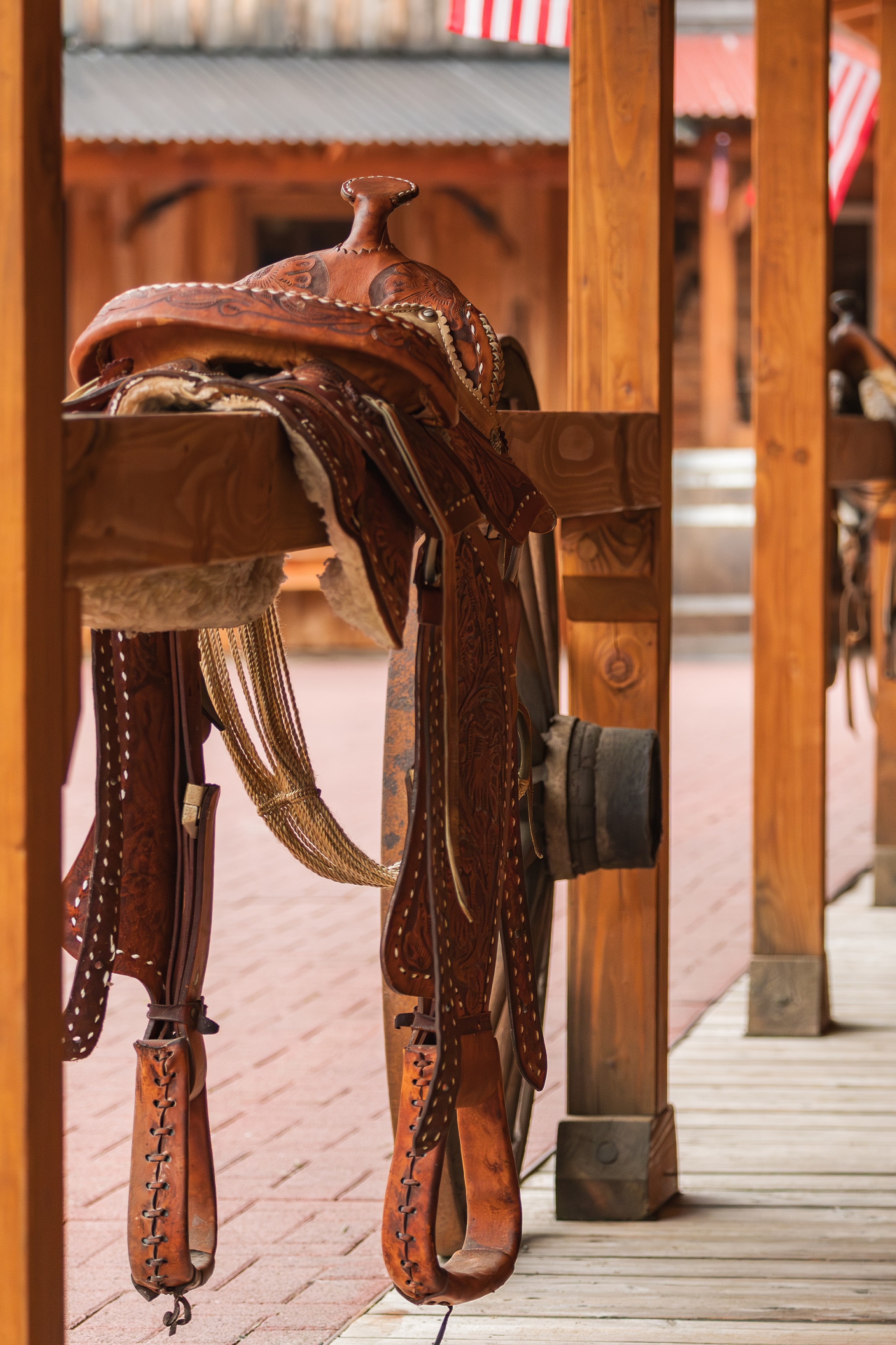 Das Bild zeigt einen Sattel, der auf einem hölzernen Sattelbock ruht. Der Sattel ist aus braunem Leder gefertigt und mit aufwendigen Mustern verziert. Die Steigbügel hängen an den Seiten des Sattels. Im Hintergrund sind Holzsäulen und eine amerikanische Flagge zu sehen. Die Szene scheint sich im Freien zu befinden, möglicherweise auf einer Ranch oder in einer Westernstadt.