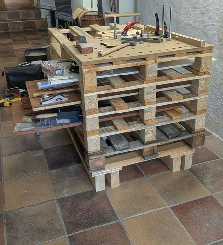 eine Werkbank aus 5 Paletten und anderem Holz, nach links ragen 3 eingeschobene Bretter raus, auf denen Werkzeug liegt. Obenauf eine Lochplatte mit Hobel und Hammer und Niederhaltern