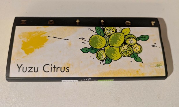 ein Foto von einer Schokoladentafelverpackung von Yuzu Citrus von Zotter. Mit einem gelben Fleck links oben und einer Traube grüner, z.T. halbierter Citursfrüchte