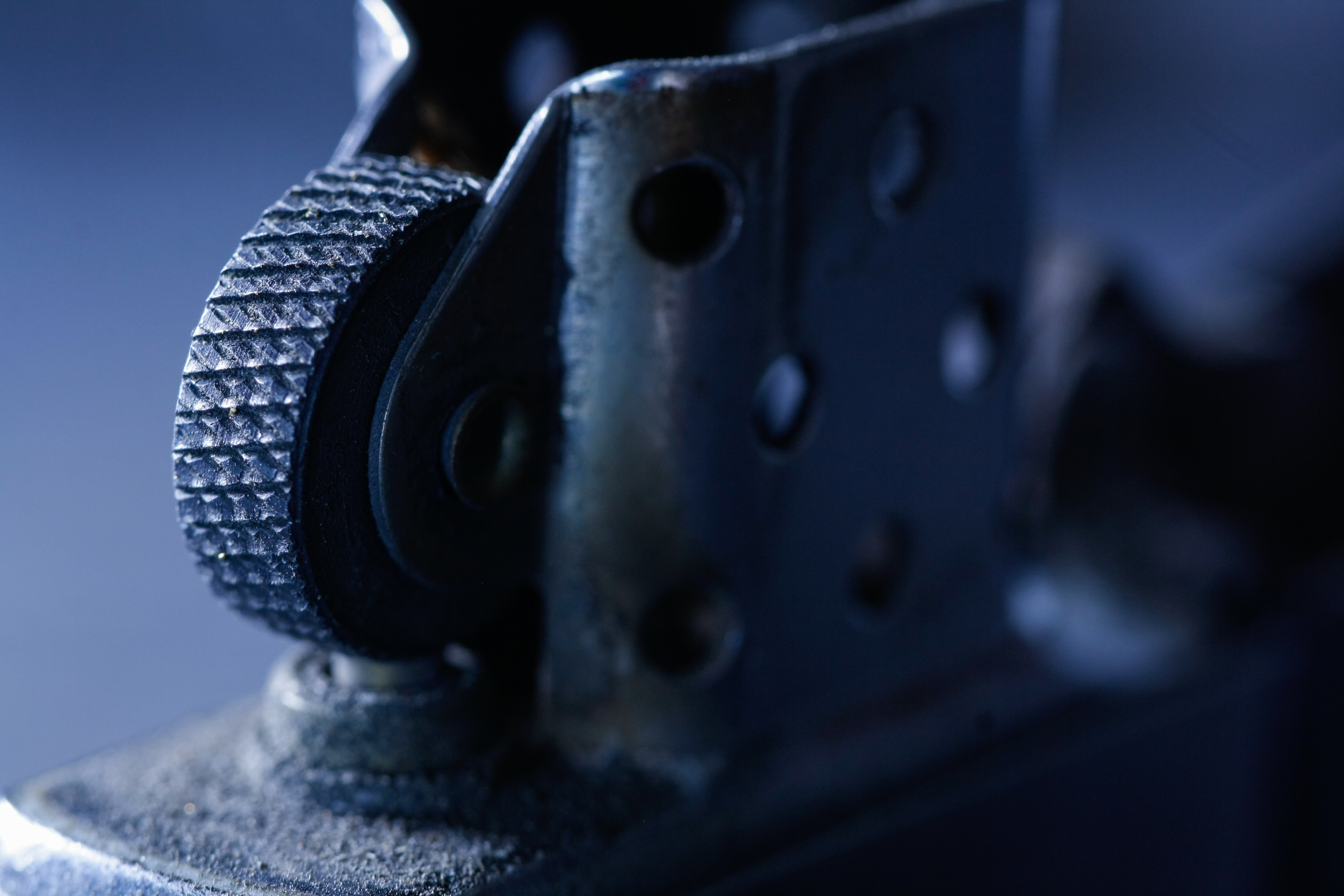 Das Bild zeigt einen Detailausschnitt eines Feuerzeugs, vermutlich vom Typ Zippo. Der Fokus liegt auf dem Reibrad und dem oberen Teil des Gehäuses. Die metallischen Oberflächen scheinen abgenutzt und weisen Gebrauchsspuren auf. Das Bild ist in einer dunklen, bläulichen Farbpalette gehalten.