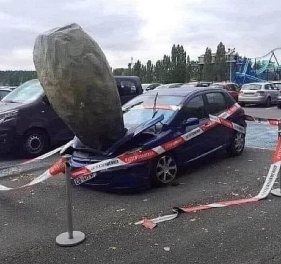 Ein kaputtes Auto steht auf einem Parkplatz. In seiner Motorhaube steckt ein Hinkelstein.