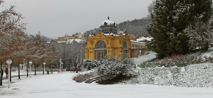 Links im Bild fängt ein geräumter Weg sich durch einen verschneiten Park zu schlängeln. Ihn säumen weiße Laternenmäste mit kugelfärmigen Schirmen darauf und einen Alllee von Bäumen links neben dem Weg. Am Ende des Wegs eine große Halle in ockergelb mit einem großen Grundbogenfenster. Rechts des Parks beginnt ein kleines Wäldchen, alles bedeckt mit einer ca. 5 cm hohen Schneeschicht.