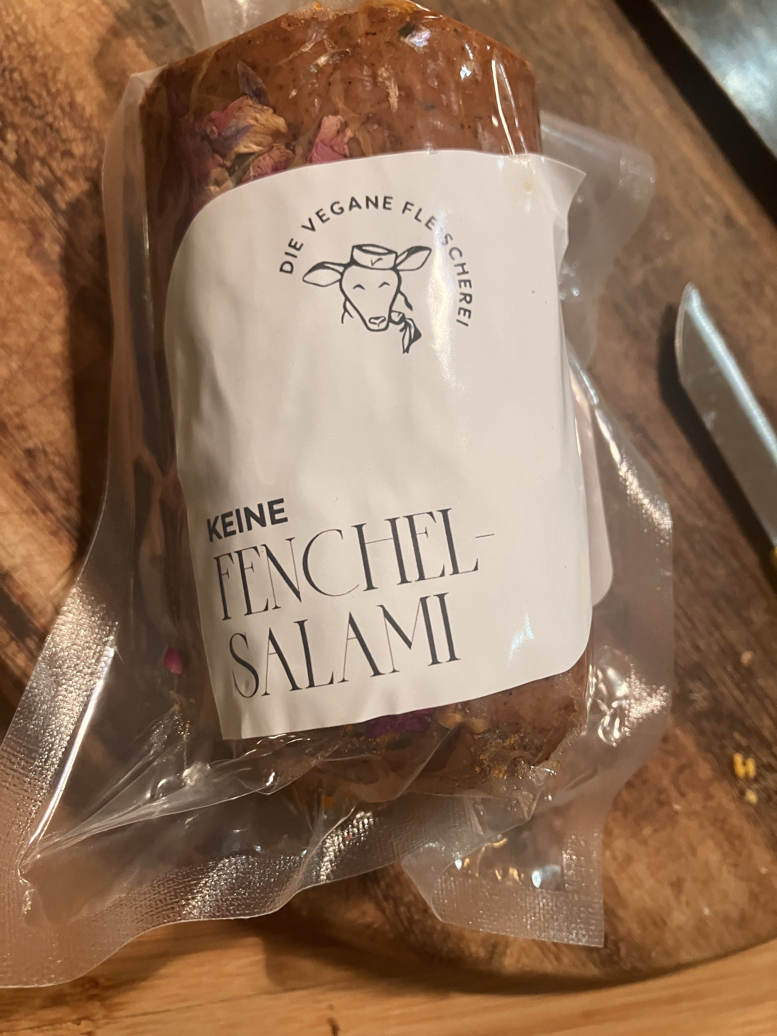 Seitansalami in einer Plastikverpackung. Auf dem schlichten Etikett steht “Die Vegane Fleischerei: Keine Fenchelsalami”.
Das Logo ist der Kopf eines Schafs mit einem Hütchen auf dem das Vegan Logo prangt. Das Schaf kaut genüsslich auf einer Karotte.