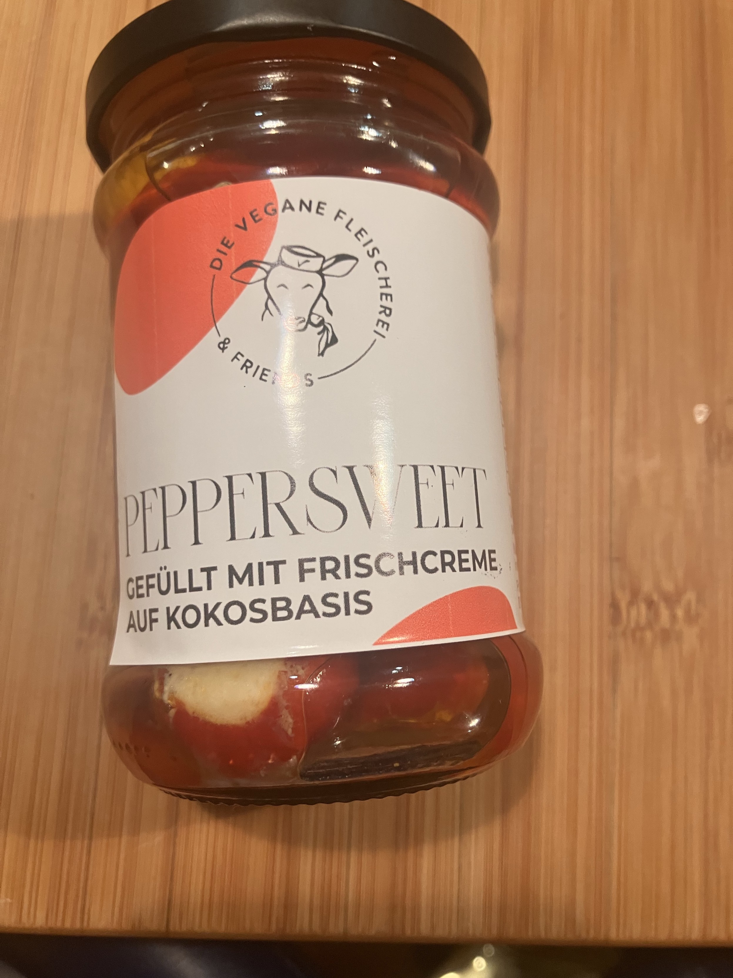 Gefüllte Kirschpaprika in Öl in einem Einmachglas. Auf dem schlichten Etikett steht “Die Vegane Fleischerei & Friends: Peppersweet gefüllt mit Frischcreme auf Kokosbasis”.
Das Logo ist der Kopf eines Schafs mit einem Hütchen auf dem das Vegan Logo prangt. Das Schaf kaut genüsslich auf einer Karotte.