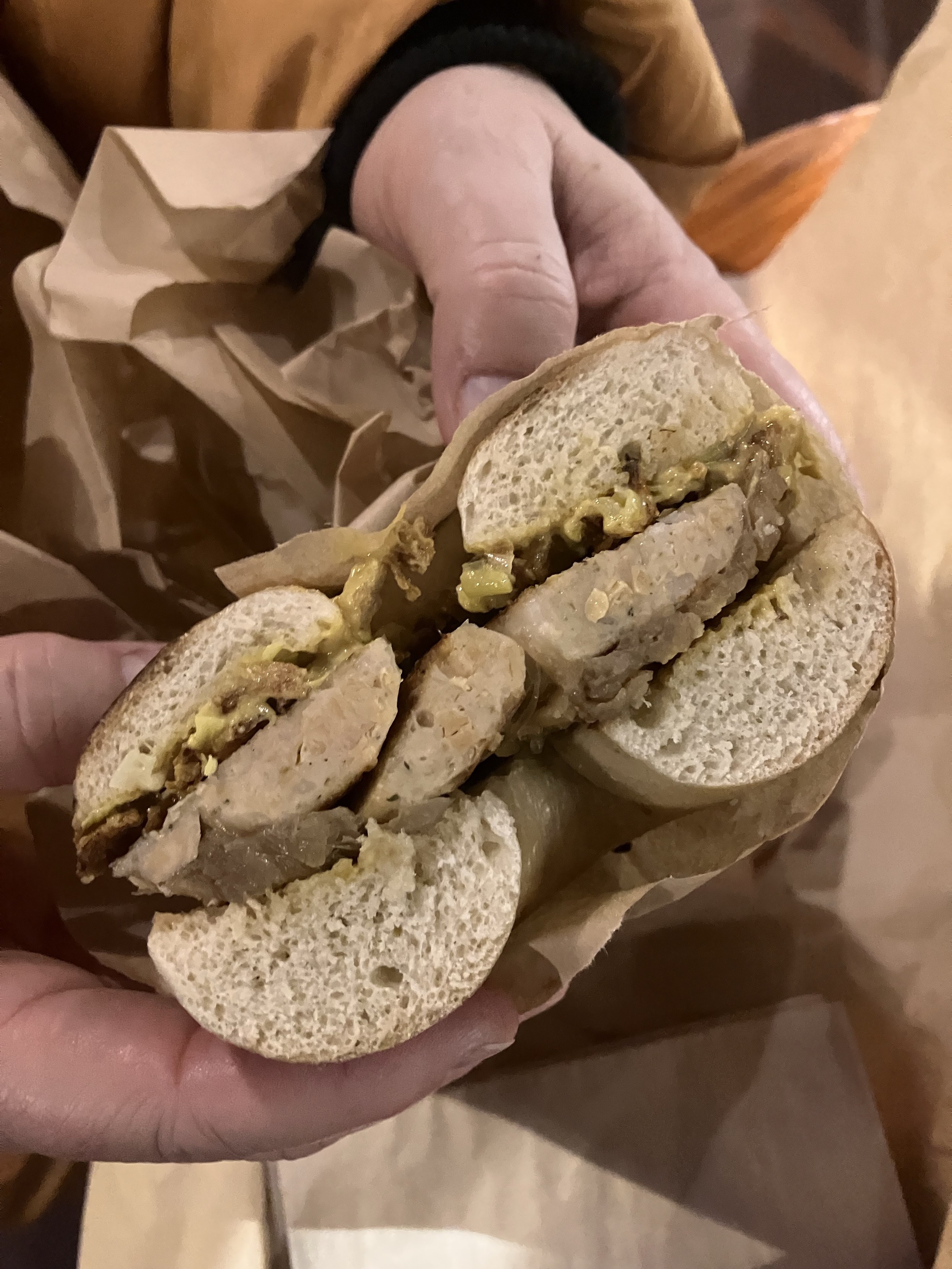 Eine Person hält die Hälfte eines Bagels mit veganer Rostbratwurst, Sauerkraut und Senf. Der Bagel ist in braunes Papier gewickelt.