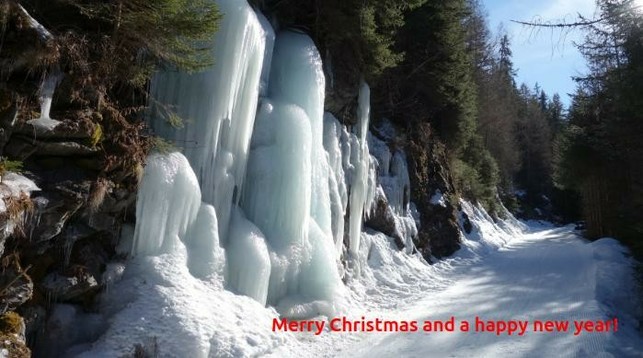 Waldweg durch verschneiten Winterwald. Links vom Weg ein gefrorener Wasserfall, dessen lange dicke Eiszapfen in der Mittagsssonne glitzernt und funkeln. Der verschneite Waldweg führt bergan und verliert sich im Nadelwald aus Fichten und Tannen. 