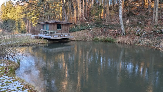 Das Bild zeigt eine kleine Hütte am Rand eines Mischwalds bestehend aus hohen Fichten und Buchenstämmen an einem kleinen Fischteich von der Fläche von 10 auf 20 Meter. Der Rand des Teichs ist mit Schneeresten bedeckt. Die Terrasse, die an der Hütte angebaut ist, ragt ein wenig über den Teich, der mit einer dünnen Eisschicht bedeckt ist. Auf der Terrasse steht eine Sitzgruppe, die Hütte hat ein Fenster zur Teichseite hin. Die Bäume des Waldes spiegeln sich auf der dünnen Eissschicht des Teichs.