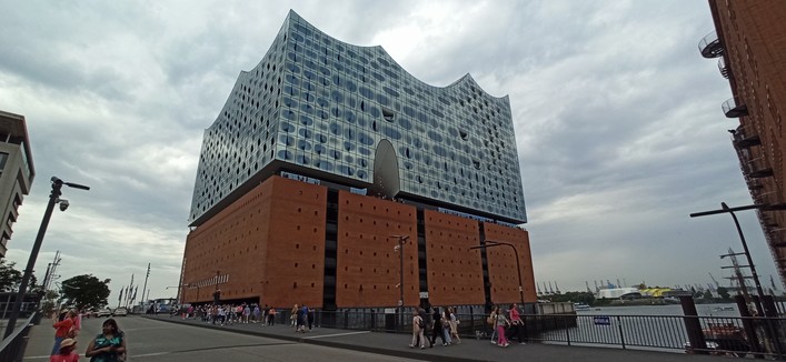Elbphilharmonie aus der seitlichen Perspektive von unten aus der Persepktive der Fußgänerbrücke aus. Die nach sußen gebogenen Glasfenster ziehen sich wie Augenlider über Augen, dabei ist die Form pro Fenster individuell in ihrer Größe. Die Glasfassade beginnt in ca. 30 Meter Höhe, darunter die alten Speicherbauten aus rotem Backstein.