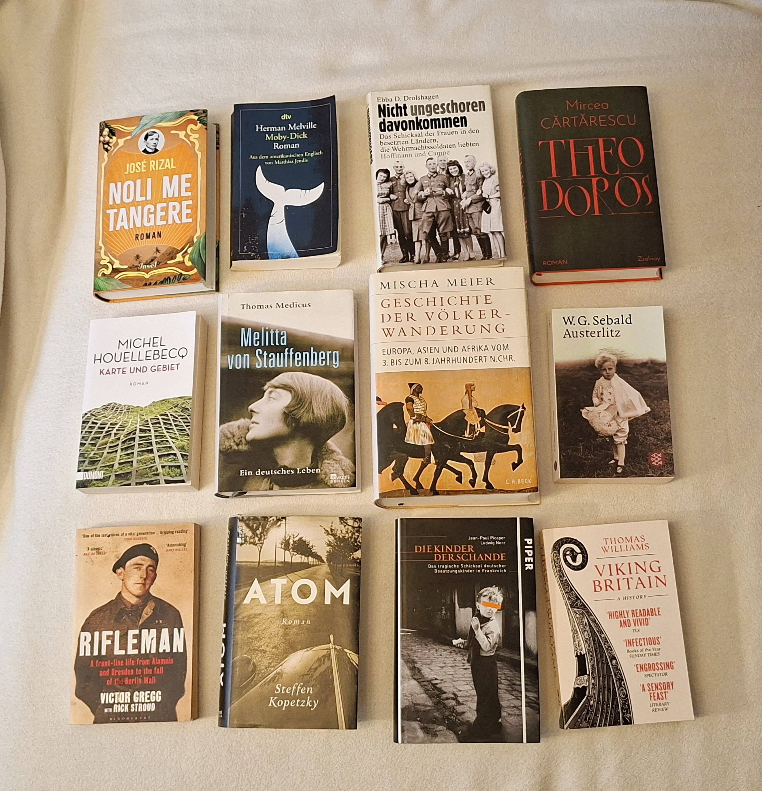 "Neun Bücher liegen ausgebreitet auf einer hellen Oberfläche. Von links oben nach rechts unten sind folgende Titel zu sehen: 'Noli me tangere' von José Rizal, 'Moby Dick' von Herman Melville, 'Nicht ungeschoren davongekommen' von Ebba D. Drolshagen, 'Theo Doros' von Mircea Cărtărescu, 'Karte und Gebiet' von Michel Houellebecq, 'Melitta von Stauffenberg' von Thomas Medicus, 'Geschichte der Völkerwanderung' von Mischa Meier, 'Austerlitz' von W.G. Sebald, 'Rifleman' von Victor Gregg mit Rick Stroud, 'Atom' von Steffen Kopetzky, 'Die Kinder der Schande' von Jean-Paul Picaper und Ludwig Norz, und 'Viking Britain' von Thomas Williams."