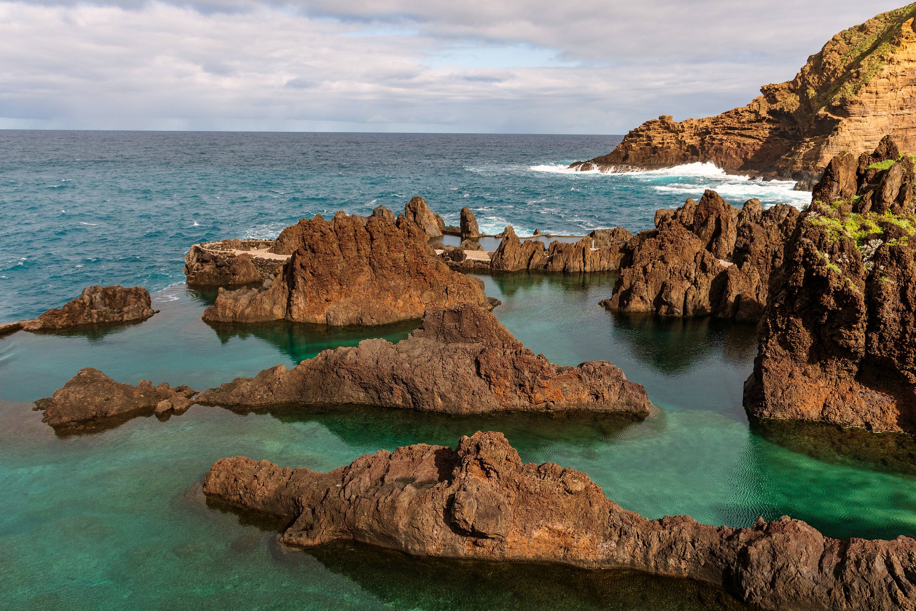 Das Bild zeigt eine Küstenlandschaft mit Felsen im Vordergrund, die von klarem, türkisblauem Wasser umgeben sind. Im Hintergrund ist der offene Ozean zu sehen, der bis zum Horizont reicht, wo er auf einen bewölkten Himmel trifft. Auf der rechten Seite des Bildes befindet sich ein felsiger Hügel, der mit Vegetation bedeckt ist. Die Felsen im Wasser bilden natürliche Pools.