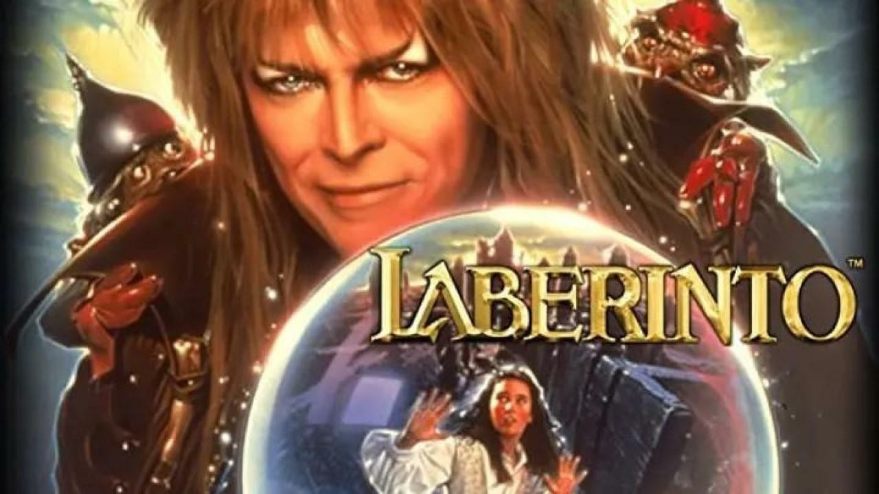 Spanish backdrop for 'Labyrinth', featuring a dramatic fantasy composition. At the top, a figure with long hair and an intense expression wears dark cloak, on his shoulders two small fantasy figures. In the center, a large crystal ball contains an image of a girl in a white nightgown with her hands raised, with a labyrinth maze visible behind her. The title 'LABERINTO' appears in ornate gold fantasy-style lettering. The background features blue and gold lighting with a mystical atmosphere.