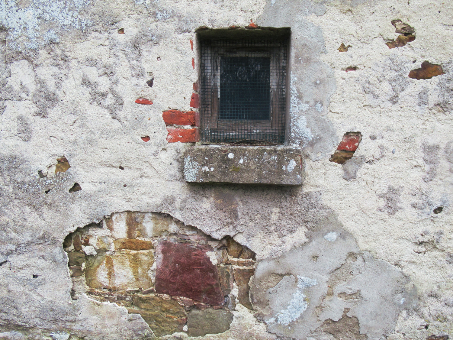 Ein Stück Fassade eines alten Gebäudes aus Bruchstein. Die ehemals weiße Fassadenfarbe ist an vielen Stellen abgebröckelt. Ungefähr in der Bildmitte ein kleines Fenster mit einem verwitterten Holzrahmen. Davor ein Gitterdraht.