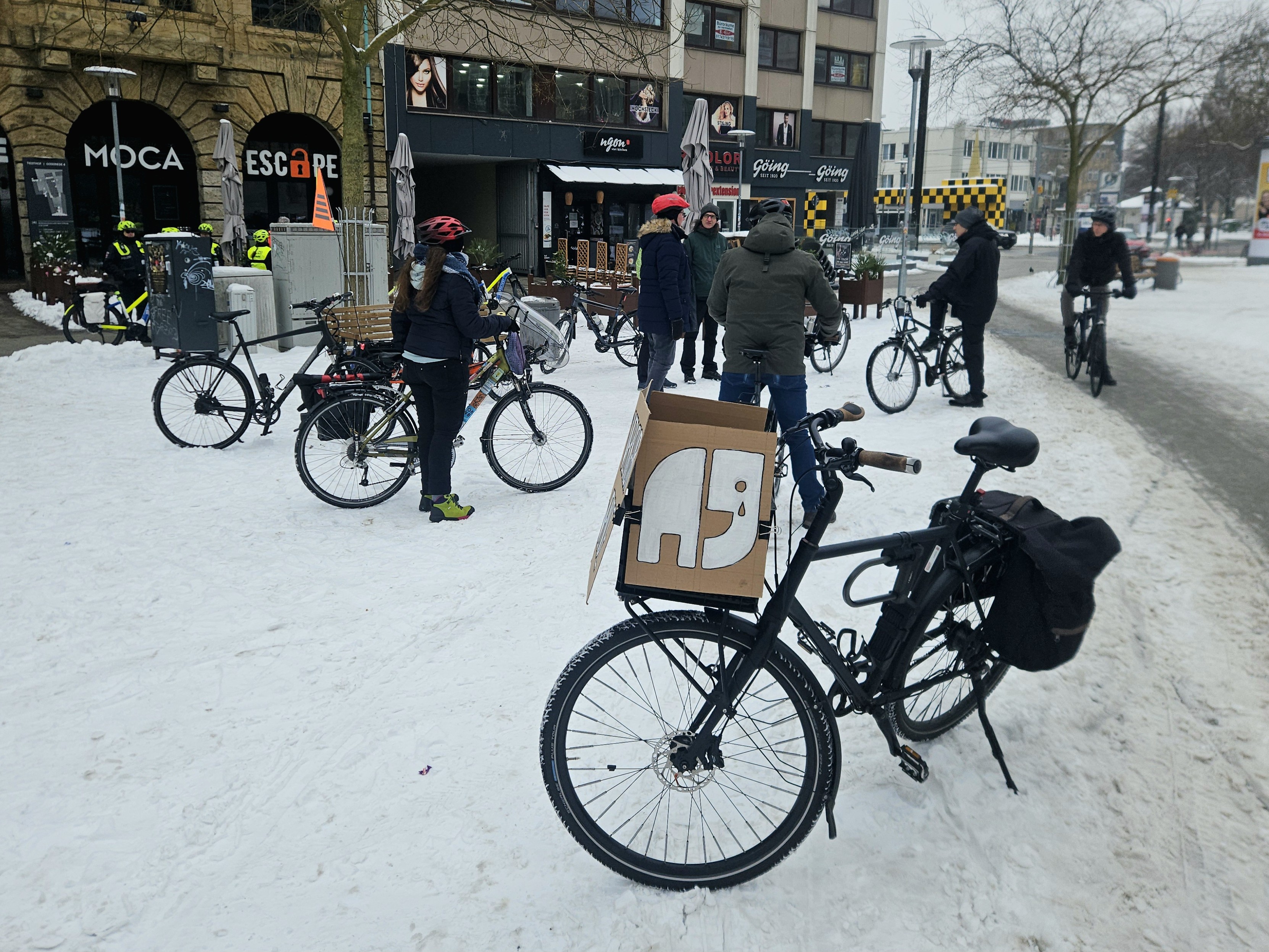 Foto einer Gruppe von Menschen und Fahrrädern auf einem von Schnee bedeckten Platz. Im Hintergrund sind noch ein paar Polizist:innen mit Fahrrädern zu sehen.