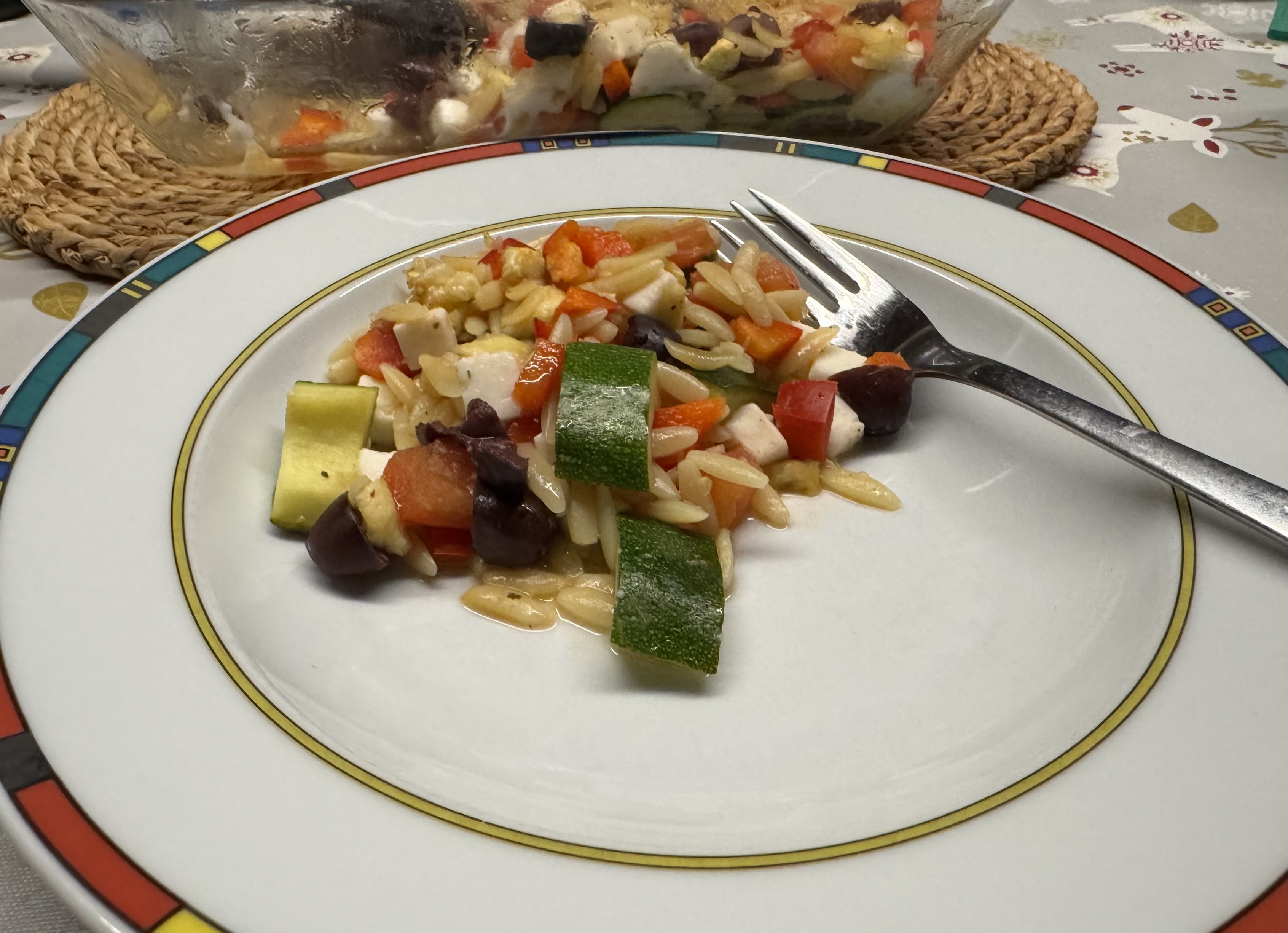 Eine kleine Portion des Zucchini-Gerichts auf einem Teller mit buntem Randdekor, am rechten Rand eine Gabel, im Hintergrund die Glasbackform auf zwei Untersetzern.