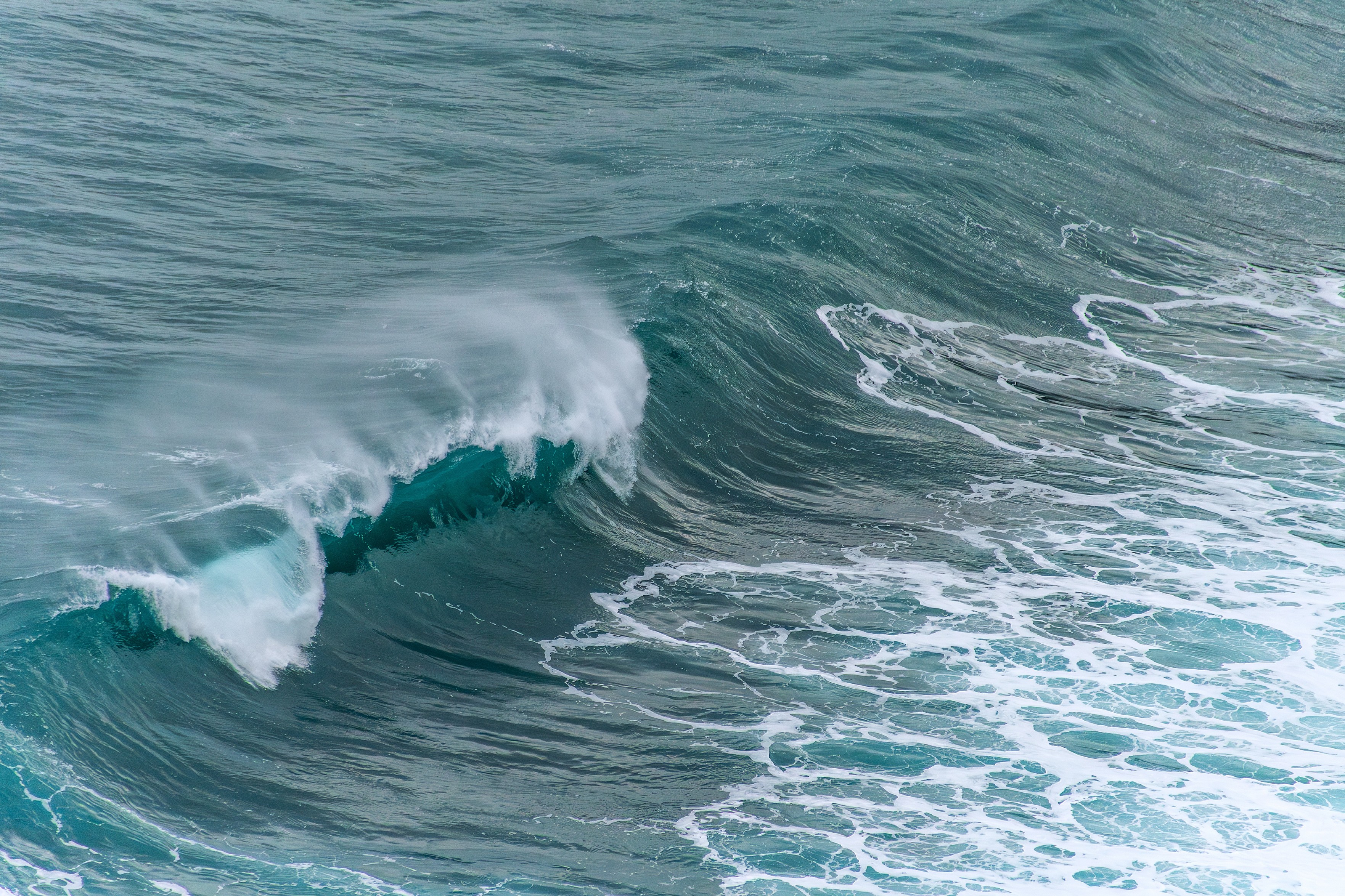 Ein Foto einer Welle, die sich im Meer bricht. Die Welle ist von einem dunklen Türkis bis zu einem helleren Aquamarin gefärbt, mit weißen Gischtkronen und Schaum. Die Bewegung des Wassers ist deutlich sichtbar.