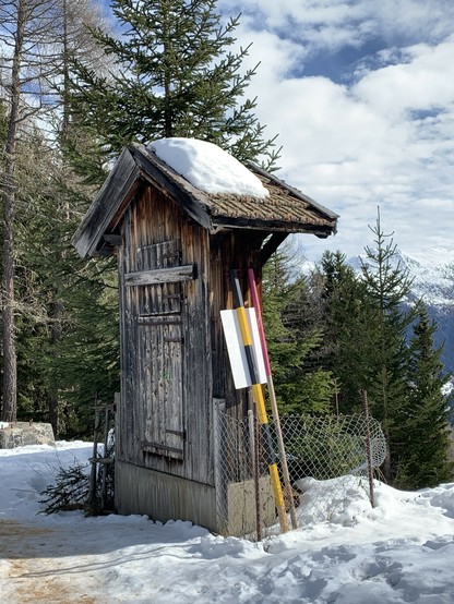 Der Wasserstandsanzeiger des Reservoirs des Berghotels Schatzalp. Die Uhr ist auf der Rückseite des wettergegerbten Holzhäuschens angebracht, welches die Mechanik schützt. Auf dem Giebeldächlein liegt wenig Schnee. Aufnahme von 2023.