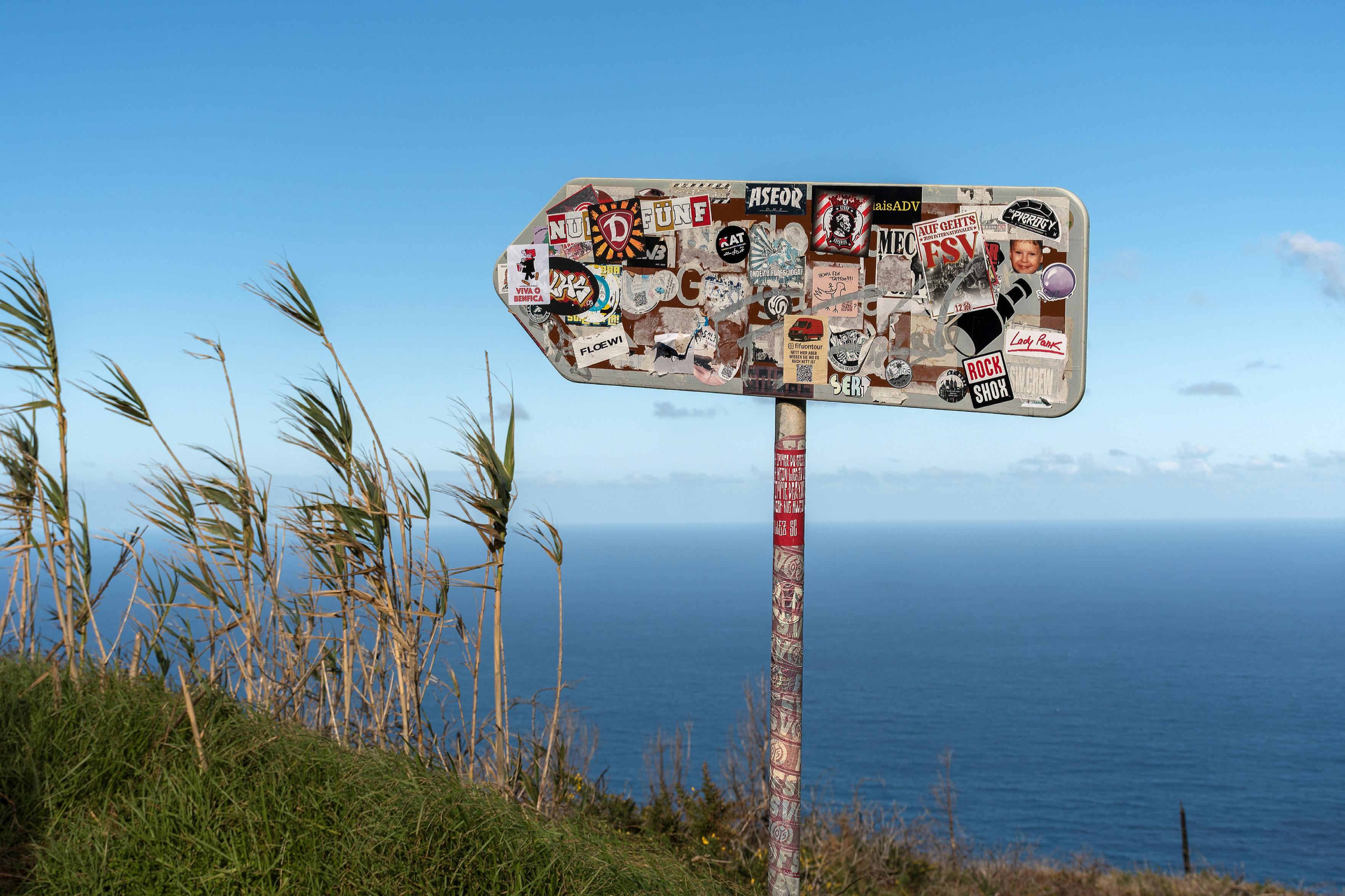 Das Bild zeigt ein mit Aufklebern bedecktes Wegweiserschild vor einer Meereskulisse an einem sonnigen Tag.
Das Foto zeigt eine Nahaufnahme eines Wegweiserschildes, das mit zahlreichen Aufklebern bedeckt ist. Das Schild ist pfeilförmig und weist nach links. Es ist an einem Pfosten befestigt, der ebenfalls mit Aufklebern und Kritzeleien versehen ist.