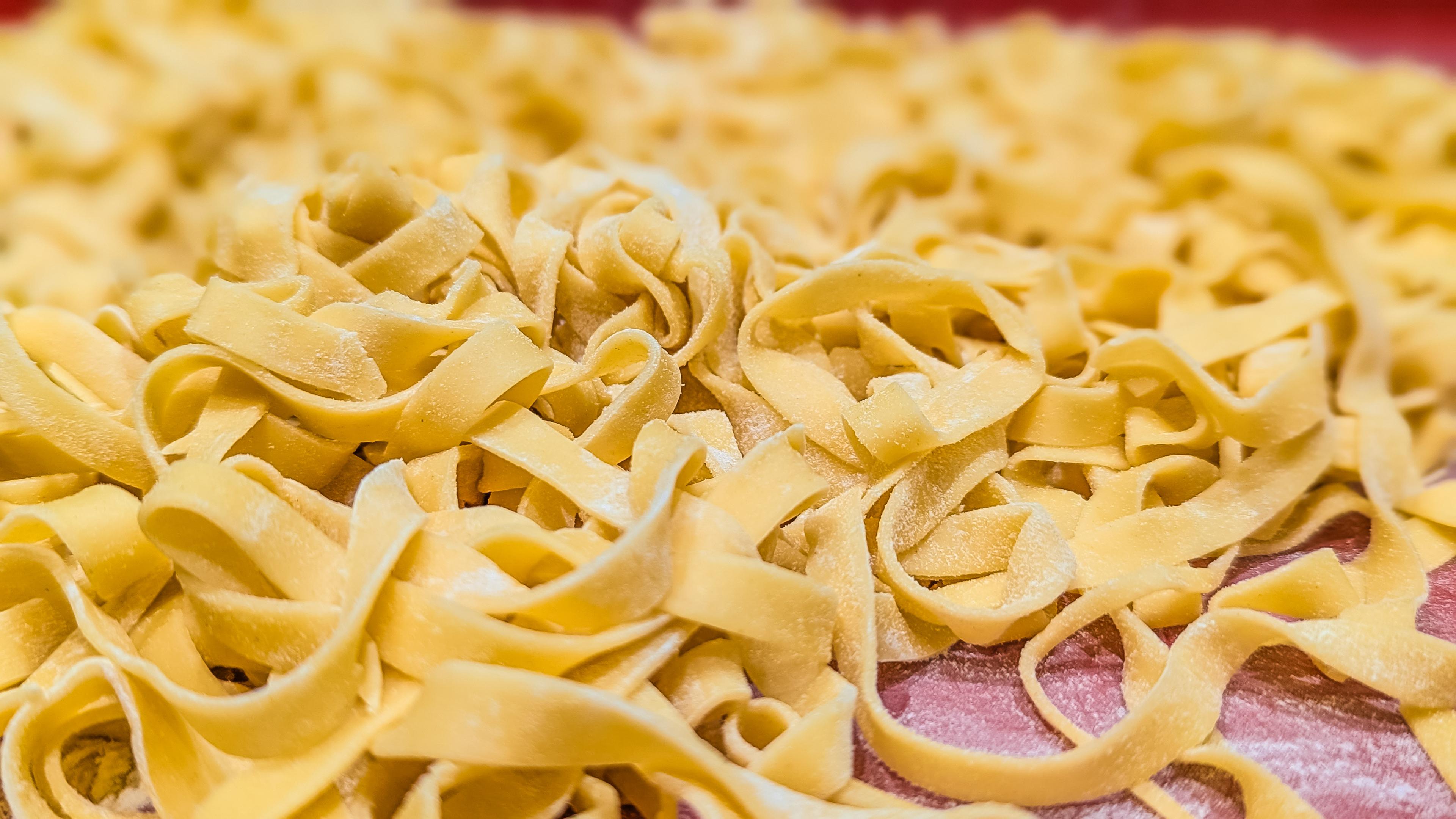 Das Bild zeigt einen appetitlichen Haufen frisch zubereiteter, handgemachter Pasta, die locker auf einer Arbeitsfläche verteilt ist. Hier sind die Details der Aufnahme:
Optik und Textur
Form: Es handelt sich um bandförmige Nudeln, vermutlich Tagliatelle oder Fettuccine. Sie sind unregelmäßig geschwungen und in lockeren Nestern oder Haufen angeordnet.
Oberfläche: Die Pasta hat eine matte, leicht raue Textur, die typisch für hausgemachten Teig ist. Man erkennt deutlich eine feine Schicht aus Mehl, die darüber gestäubt wurde, um ein Zusammenkleben zu verhindern.
Farbe: Die Nudeln haben einen warmen, goldgelben Farbton, was auf einen hohen Anteil an frischen Eigelban deutet.