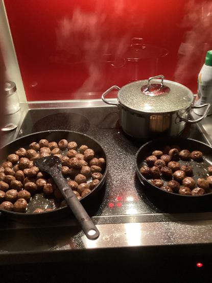 Blick auf den Glaskeramikherd. Vorn eine grosse und eine kleine Bratpfanne. Darin braten Hackfleischbällchen. Die Oberfläche des Herds ist mit Fett verspritzt.