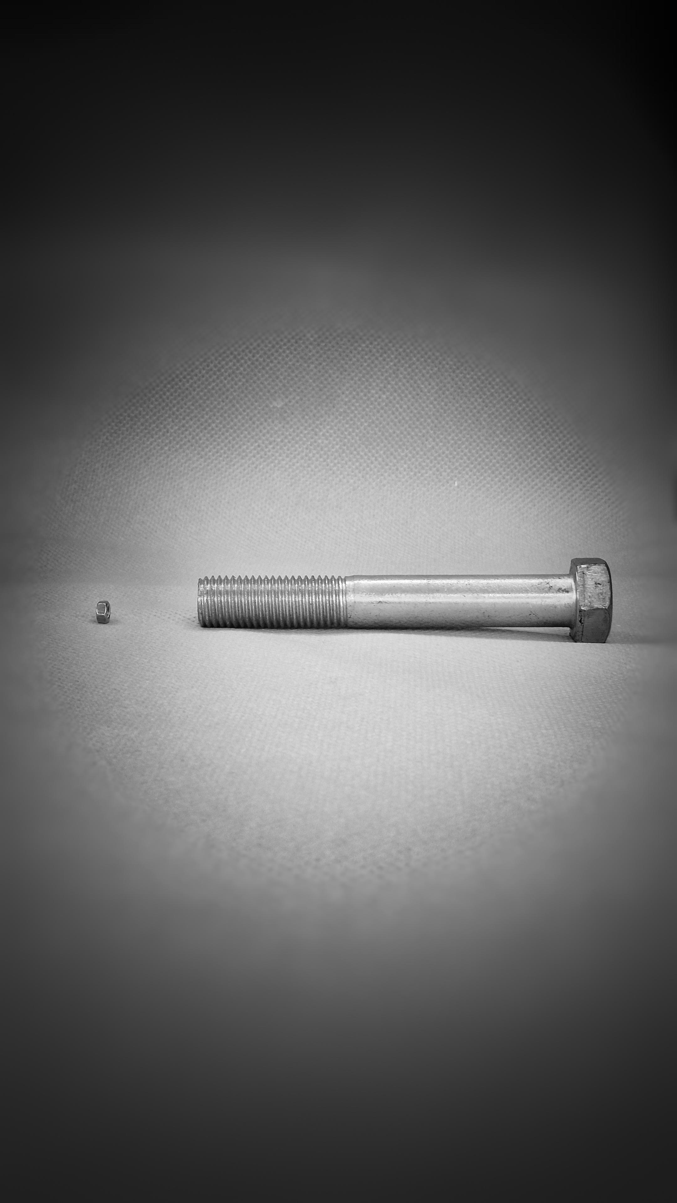 Die Schwarzweißaufnahme zeigt eine Metallschraube mit Metallgewinde und Sechskantkopf sowie eine Metallmutter. Die Aufnahme wurde von der Seite gemacht, die Schraube befindet sich auf der rechten Seite des Bildes und die Mutter ist auf der linken Seite zu sehen. Die Schraube zeigt mit ihrem Sechskantkopf nach rechts und ihrem Gewinde in Richtung Mitte des Bildes. Links im Bild ist die Mutter erkennbar, die sich mit einem gewissen Abstand zum Gewinde der Schraube befindet. Die Schraube weist eine überdurchschnittliche Größe auf, während die Mutter eine sehr kleine Größe aufweist. Die Konturen des Bildes erscheinen verschwommen.