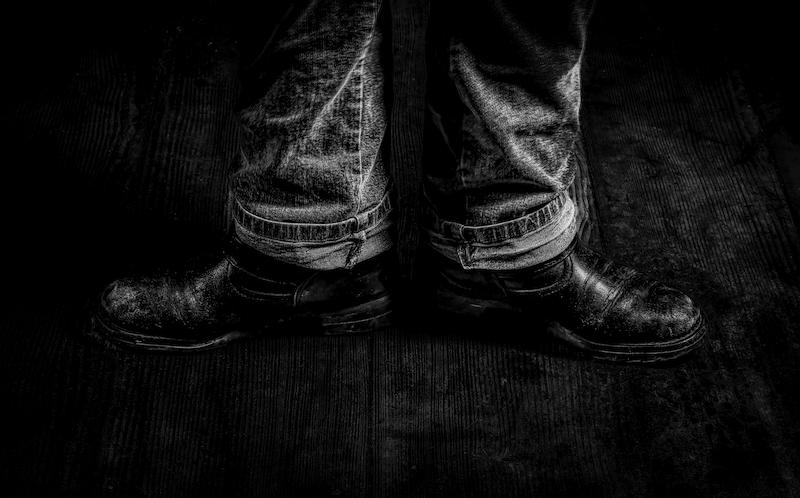 Zwei Stiefel mit dem Anfang einer Jeans des Trägers. Beide Stiefel zeigen in die entgegengesetzten Richtung. Sie stehen auf einem alten Holzboden. Das Foto ist in Schwarzweiß und ziemlich dunkel gehalten.