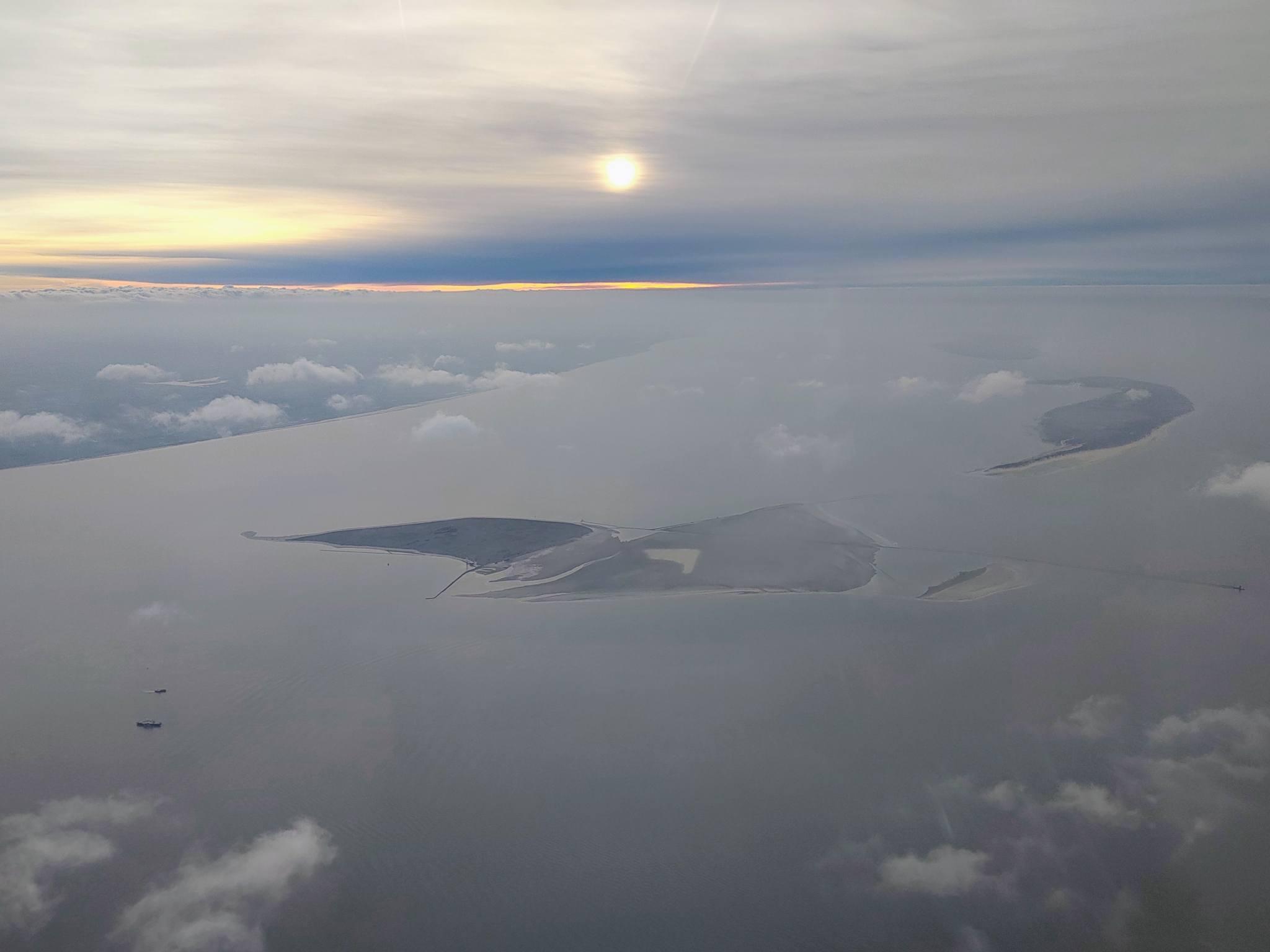 Islands Mellum and Wangerooge seen from 5000 ft. Below are small cloudlets, the sun's disk ist visible behind a hazy sky.