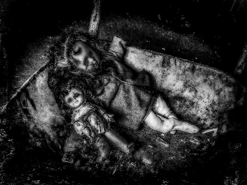 Zwei Puppen liegen in einer alten Schubkarre. Sie sehen alt und völlig runtergekommen aus. Insgesamt ein eher gruseliger Anblick. Das Foto ist in Schwarzweiß.