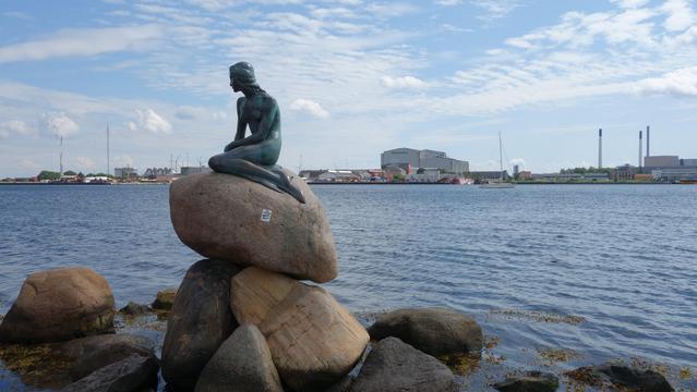 Die kleine Meerjungrau als Bronzefigur auf einem Felsen sitzend in der Sonne des Nachmittagslichts von Westen her angestrahlt, dahinter das Wasser des Kopenhageners Hafen in blau. Leicht bewölkter Himmel und Gebäude an der anderen Seite des Hafens.